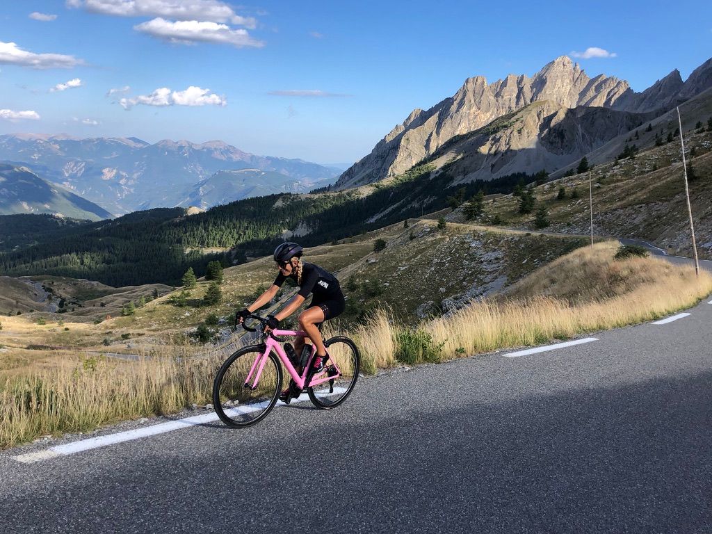 Anne à vélo sur une route de col dans les Alpes, sommets déchiquetés en arrière-plan